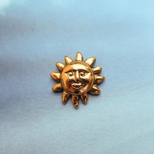 May include: A small, gold-colored sun-shaped charm with a smiling face. The charm has a detailed design with rays extending outwards. The background is a light blue color with a cloudy pattern.