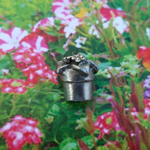 May include: A silver charm in the shape of a flower pot with a flower on top. The charm is sitting on a background of pink and purple flowers.