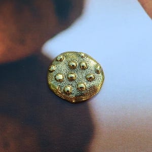 May include: A small, round, gold-colored button with a textured surface and raised, spherical details. The button is centered on a light background, with blurred, brown, textured shapes in the background. This button could be used for clothing or crafts.