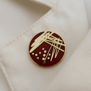 May include: A red and gold enamel pin with a white design of lines and dots. The pin is circular and has a gold metal backing.
