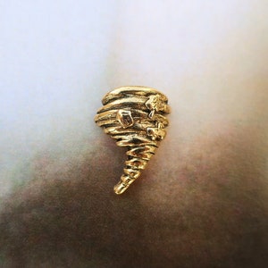 May include: A gold-toned metal pin shaped like a tornado with a swirling design. The pin has a small, detailed design of a house at the top.