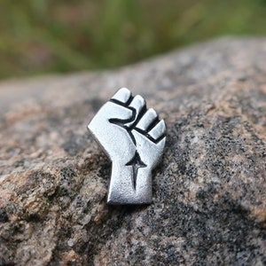 May include: A silver-coloured metal pin shaped like a raised fist with a star in the palm. The pin is sitting on a grey rock.