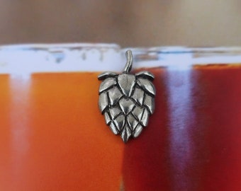 May include: A silver-colored metal hop cone charm. The charm is in focus and is in front of a blurred background of three glasses of beer.