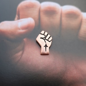 Può includere: Un pugno alzato, simbolo di forza e solidarietà, è rappresentato in metallo color rame. Il pugno è chiuso con il pollice esteso, e i dettagli delle nocche e delle dita sono chiaramente visibili.