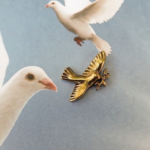May include: A gold-colored metal dove brooch, with wings spread, against a background of two white doves in flight.