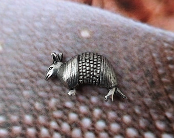 Könnte beinhalten: Eine silberne Gürtelspange in Form eines Gürteltiers mit einer detaillierten, strukturierten Schale. Die Spange befindet sich auf einem braunen Strickstoff.