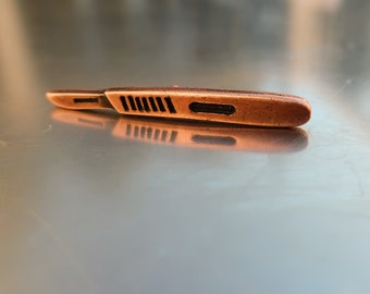 May include: A small, copper-colored scalpel lies on a reflective, metallic surface. The scalpel has a blade and a handle with a series of parallel grooves. The image is a close-up, highlighting the tool's details and texture.