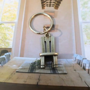 May include: A gold-toned metal keychain shaped like a chair. The chair has a simple design with a back, seat, and four legs. The keychain is hanging from a metal ring.