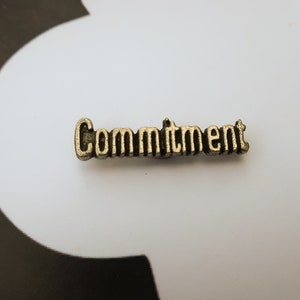 May include: A gold-colored metal charm with the word "Commitment" embossed on it.