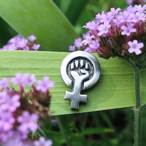 This pewter lapel pin is a raised fist inside the female symbol.  It is placed in a wild flower garden stuck through a tiger lily stalk next to purple wild flowers.