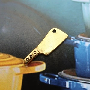 May include: A gold-colored metal charm shaped like a butcher knife. The charm is sitting on a table with a yellow and blue pot in the background.