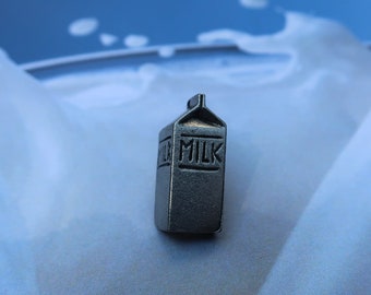 May include: A miniature silver milk carton with the word "MILK" embossed on the front, sitting on a white surface with a blue background.