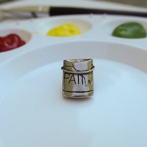Puede incluir: Un charm de plata en forma de bote de pintura con la palabra "PAINT" grabada en el lateral. El charm está sobre una paleta de pintura blanca con varios colores de pintura.