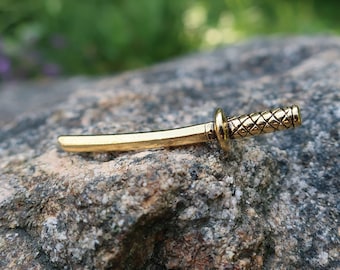 Könnte beinhalten: Ein goldfarbenes Metallsäbel mit einem strukturierten Griff. Das Säbel liegt auf einem grauen Stein.