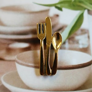 Puede incluir: Juego de cubiertos de metal dorado, que incluye un tenedor, un cuchillo y una cuchara, dispuestos verticalmente en un plato de cerámica blanco.