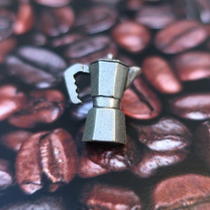 May include: A silver metal miniature coffee pot sitting on a bed of coffee beans.
