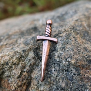 May include: A copper-colored dagger pendant with a twisted handle. The pendant is sitting on a rough, gray rock.