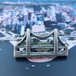 May include: A silver metal bridge charm with a detailed design. The charm is sitting on a photo of a city skyline.