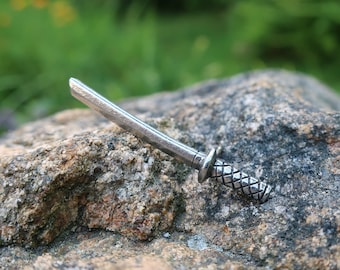 Könnte beinhalten: Ein silberfarbenes Miniatur-Katana-Schwert mit einem schwarz-weißen gemusterten Griff. Das Schwert liegt auf einem grauen Stein.