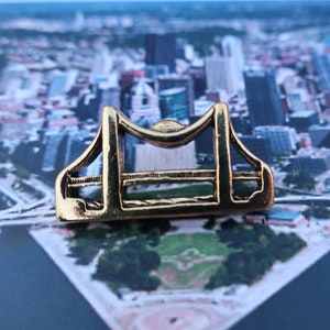 May include: A gold-colored metal bridge pin with a detailed design, sitting on a postcard image of a city skyline.