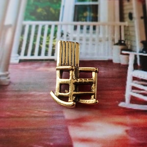 Pode incluir: Um amuleto de cadeira de balan&ccedil;o de metal dourado. O amuleto &eacute; uma r&eacute;plica em miniatura de uma cadeira de balan&ccedil;o tradicional. O amuleto est&aacute; sentado em uma superf&iacute;cie com padr&atilde;o vermelho e branco.