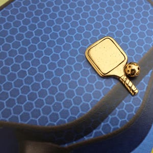 May include: A gold-colored metal charm in the shape of a pickleball paddle and ball. The paddle is flat and has a textured surface. The ball is round and has a dimpled surface.