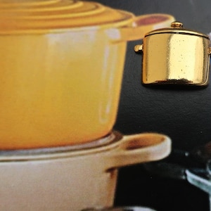 May include: A gold-colored miniature pot with a lid and handles. The pot is sitting on a table with other pots in the background.