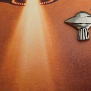 May include: A silver-colored metal pin shaped like a flying saucer. The pin is on a brown background with two blurry images of flying saucers in the background.