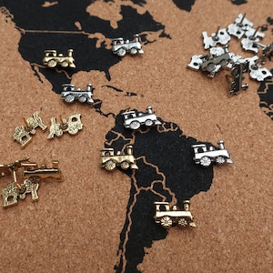 May include: A corkboard with a world map printed on it.  There are several silver and gold train-shaped push pins scattered across the map.