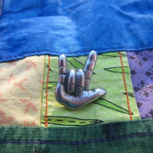 May include: A silver-colored metal pin shaped like a hand signing the American Sign Language symbol for "I love you". The pin is on a multi-colored patchwork fabric background.