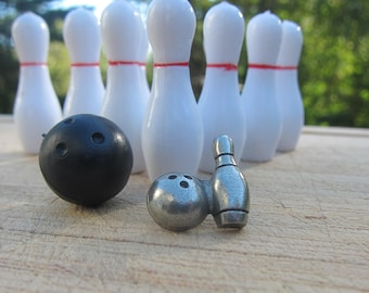 May include: A set of white bowling pins with red stripes, a black bowling ball, and two silver miniature bowling pins.