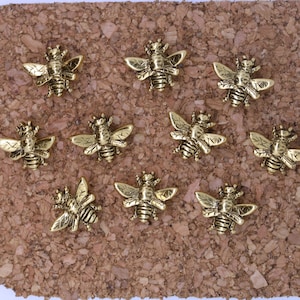 May include: Ten gold bee push pins on a corkboard background. The bees are detailed with wings, legs, and antennae.