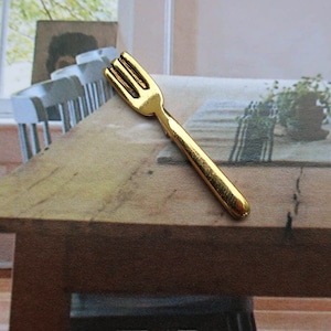Könnte beinhalten: Eine kleine, goldfarbene Gabel mit drei Zinken liegt auf einem Holztisch. Die Gabel hat eine glatte, glänzende Oberfläche. Der Tisch besteht aus hellbraunem Holz und hat ein rustikales Aussehen. Der Hintergrund umfasst ein Fenster und Stühle.
