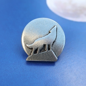 May include: A silver-toned pin featuring a wolf howling at the moon. The wolf is silhouetted against a circular background.