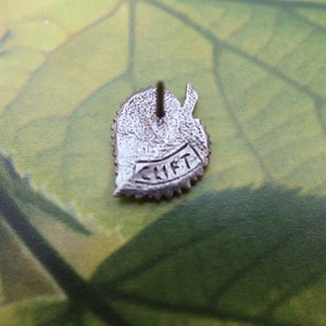 May include: A silver-colored leaf-shaped pin with the word "CLIFT" inscribed on a banner. The pin has a textured surface and a pin backing. The background is a green and brown leafy pattern.