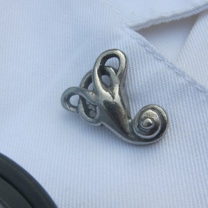 Puede incluir: Un broche de nudo celta de plata con un diseño de espiral. El broche está sobre un fondo de tela blanca.