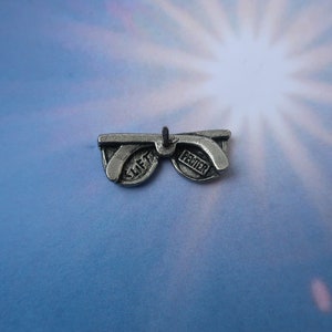 Puede incluir: Encanto de plata en forma de gafas de sol con las palabras "Slift" y "Pewter" en las lentes.