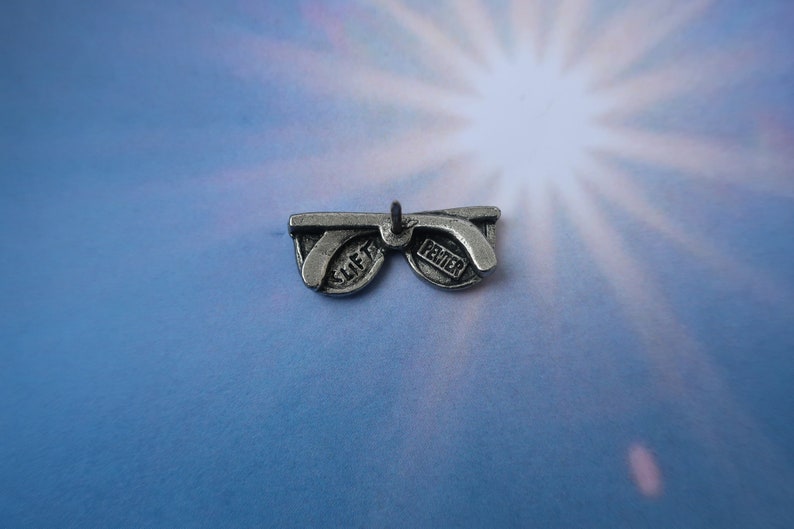 Puede incluir: Encanto de plata en forma de gafas de sol con las palabras "Slift" y "Pewter" en las lentes.