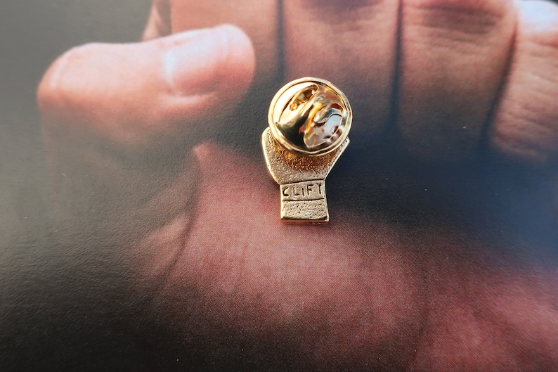 May include: Gold-colored boxing glove pin with the word "CLIFF" engraved on the glove. The pin has a safety clasp on the back.