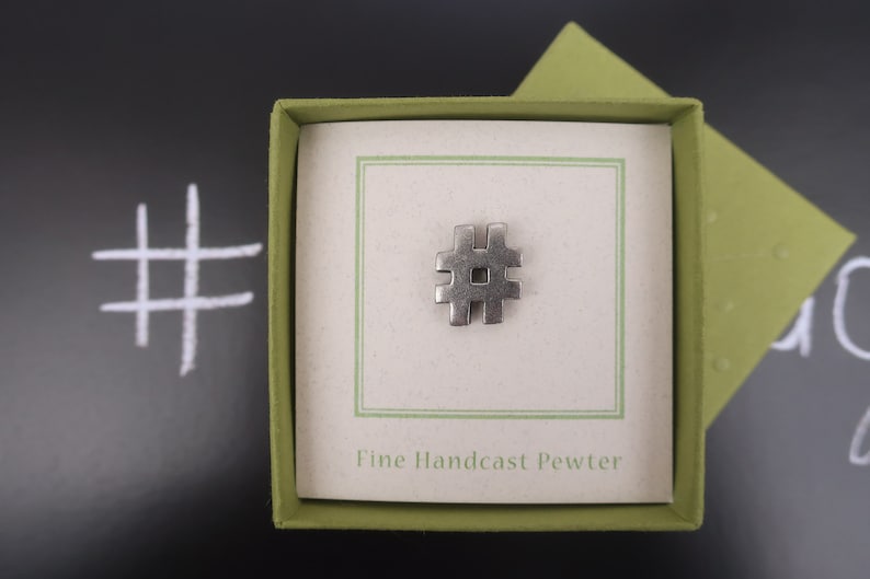 May include: A silver pewter hashtag charm in a green box with the text "Fine Handcast Pewter".