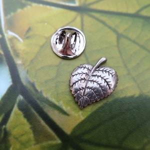 May include: A silver-colored leaf-shaped lapel pin with detailed veining, next to its round backing. The pin is on a green and yellow background, suggesting a nature-themed accessory.