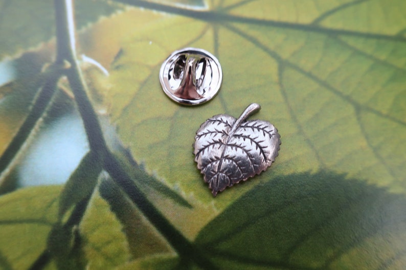 May include: A silver-colored leaf-shaped lapel pin with detailed veining, next to its round backing. The pin is on a green and yellow background, suggesting a nature-themed accessory.