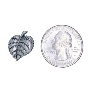 May include: A small, silver-colored leaf-shaped charm with detailed veining. Next to it is a U.S. quarter coin for size comparison. The charm is likely made of metal and has a slightly textured surface.