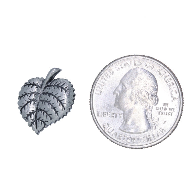 May include: A small, silver-colored leaf-shaped charm with detailed veining. Next to it is a U.S. quarter coin for size comparison. The charm is likely made of metal and has a slightly textured surface.