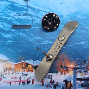 Puede incluir: Pin de metal plateado de una tabla de snowboard con la palabra "CLIFT" en ella. El pin est&aacute; sujeto a una imagen de fondo de una estaci&oacute;n de esqu&iacute; con un remonte.