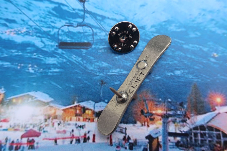 Puede incluir: Pin de metal plateado de una tabla de snowboard con la palabra "CLIFT" en ella. El pin est&aacute; sujeto a una imagen de fondo de una estaci&oacute;n de esqu&iacute; con un remonte.