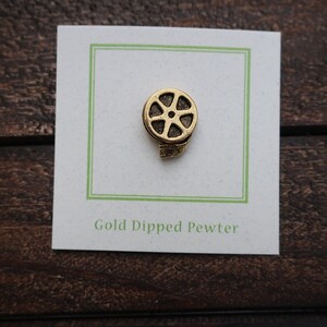 May include: Gold-dipped pewter film reel pin. The pin is on a white card with a green border and the text "Gold Dipped Pewter".