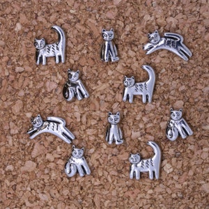 May include: Ten silver metal cat figurines in various poses, including sitting, standing, and jumping. The figurines are arranged on a corkboard background.