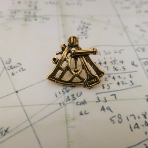 May include: A gold-colored metal sextant, a navigational tool used to measure the angle between two objects, such as a star and the horizon. The sextant is lying on a piece of paper with handwritten calculations.