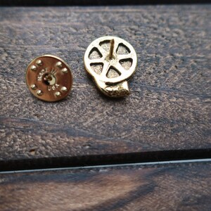 May include: Gold-toned metal film reel pin with a pin back. The back of the pin has the words "Made in USA" on it.